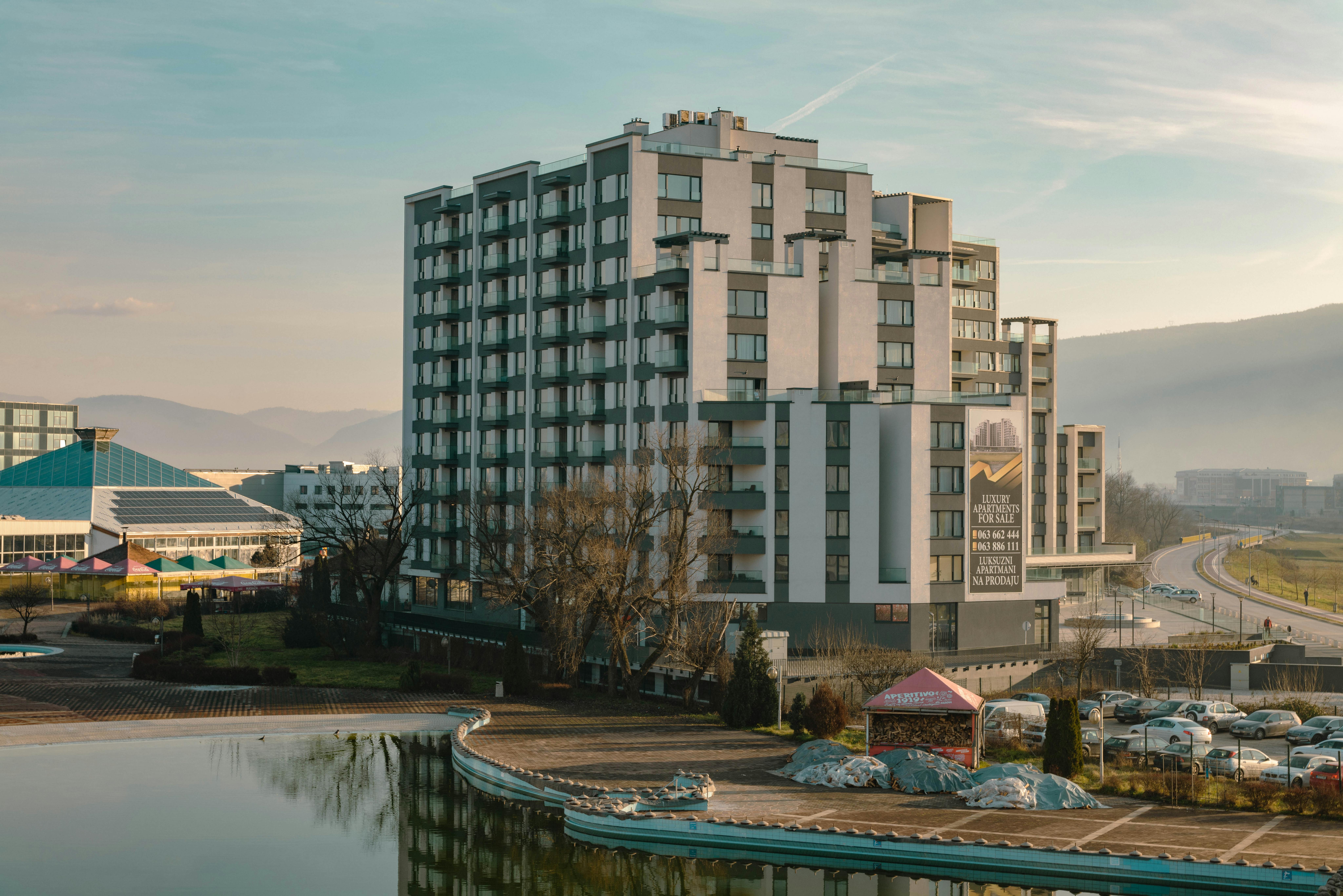 Explore a modern residential building with a serene pond and scenic mountain backdrop, ideal for urban living.
